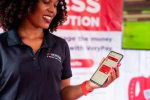 Judith, a verypay team member smiling and holding a mobile phone with the app on the screen and signage behind her